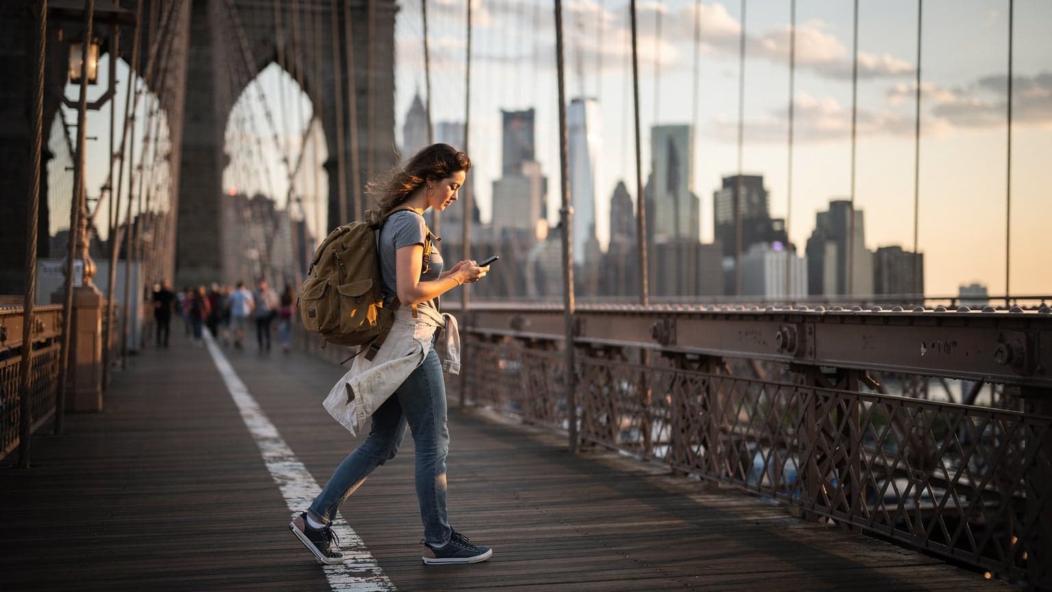 Mladá žena s batohem prochází po Brooklynském mostě v New Yorku za soumraku, drží telefon a kontroluje mapu, v pozadí panorama Manhattanu.