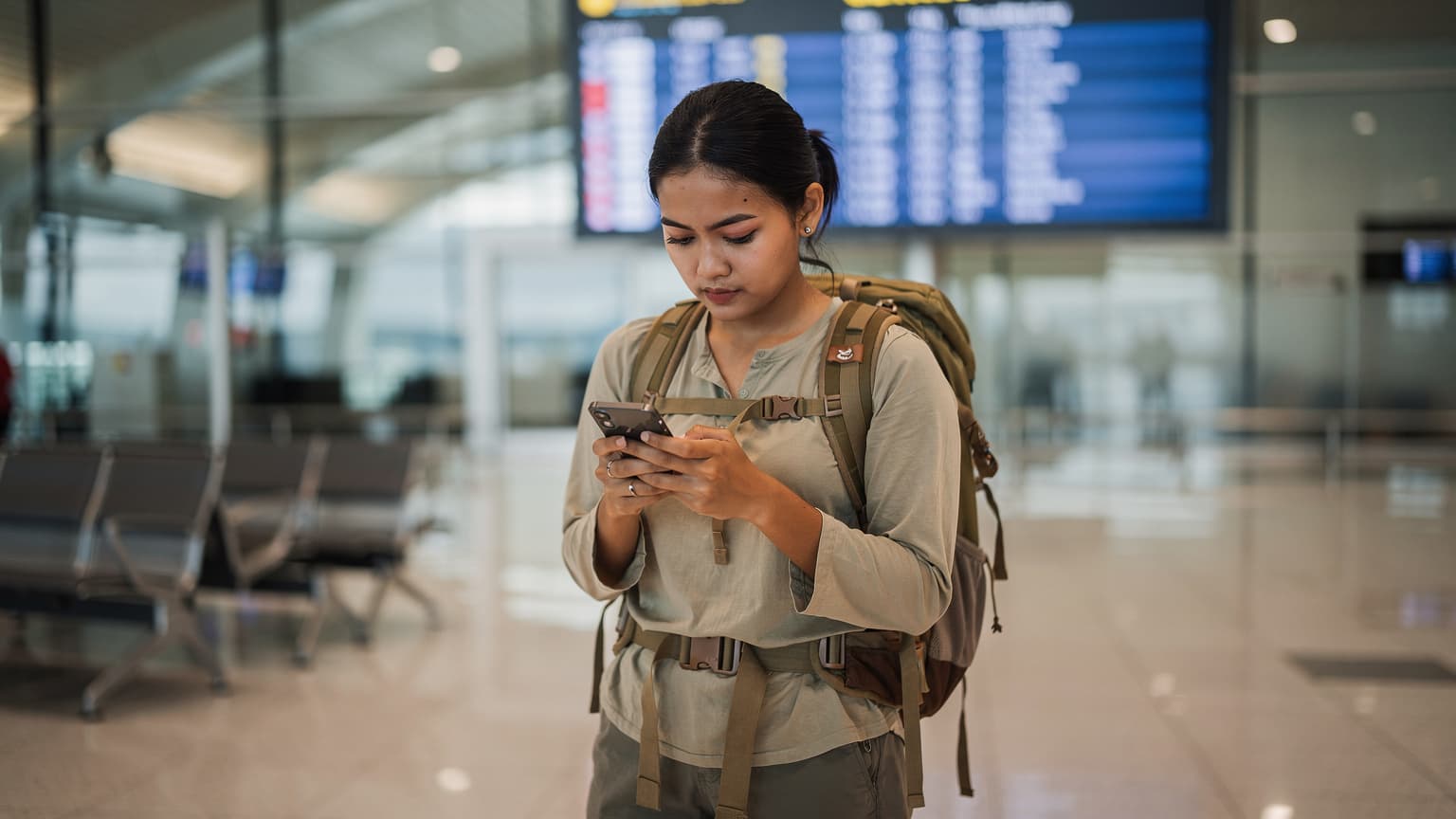 Seorang backpacker muda perempuan dari Indonesia sedang memeriksa ponselnya di Bandara Changi Singapura, dengan tas ransel besar di sampingnya dan papan keberangkatan di latar belakang, mencari sinyal atau membandingkan paket eSIM.