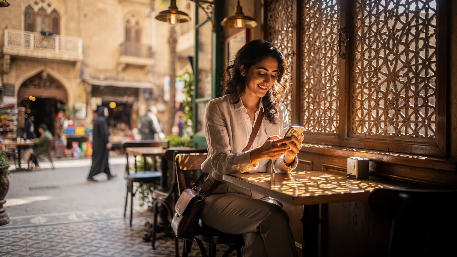 سيدة عربية شابة تبتسم وهي تحمل هاتفها الذكي وتتصفح الإنترنت أمام نافذة مقهى في منطقة الجمالية بالقاهرة، تظهر ملامح العمارة الإسلامية القديمة في الخلفية.
