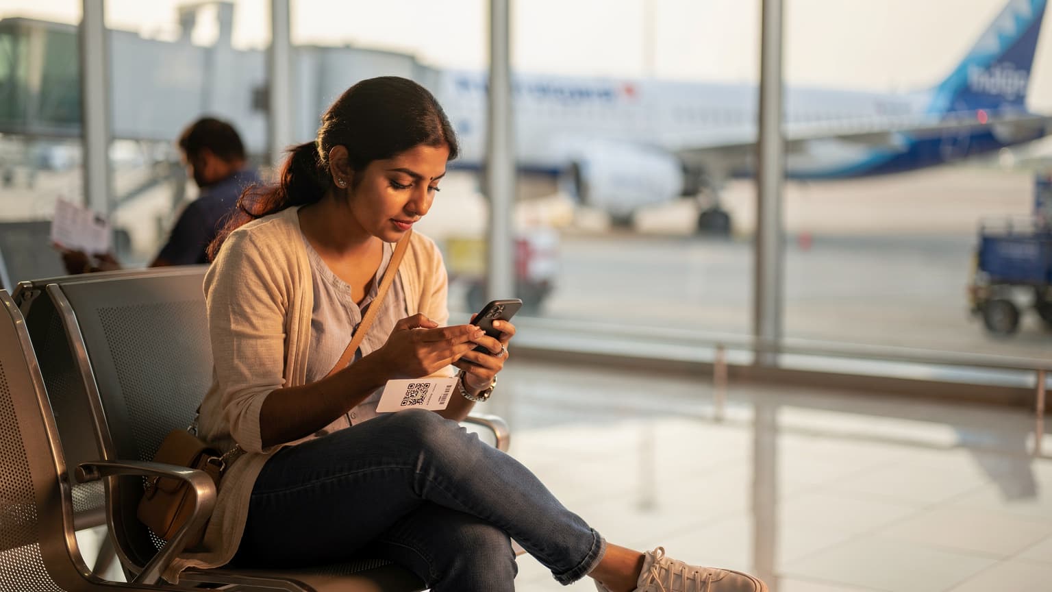 एक महिला यात्री बेंगलुरु के केम्पेगौड़ा अंतर्राष्ट्रीय हवाई अड्डे पर अपने iPhone पर eSIM एक्टिवेट कर रही है, उसके पीछे एक इंडिगो एयरलाइंस का विमान दिख रहा है।