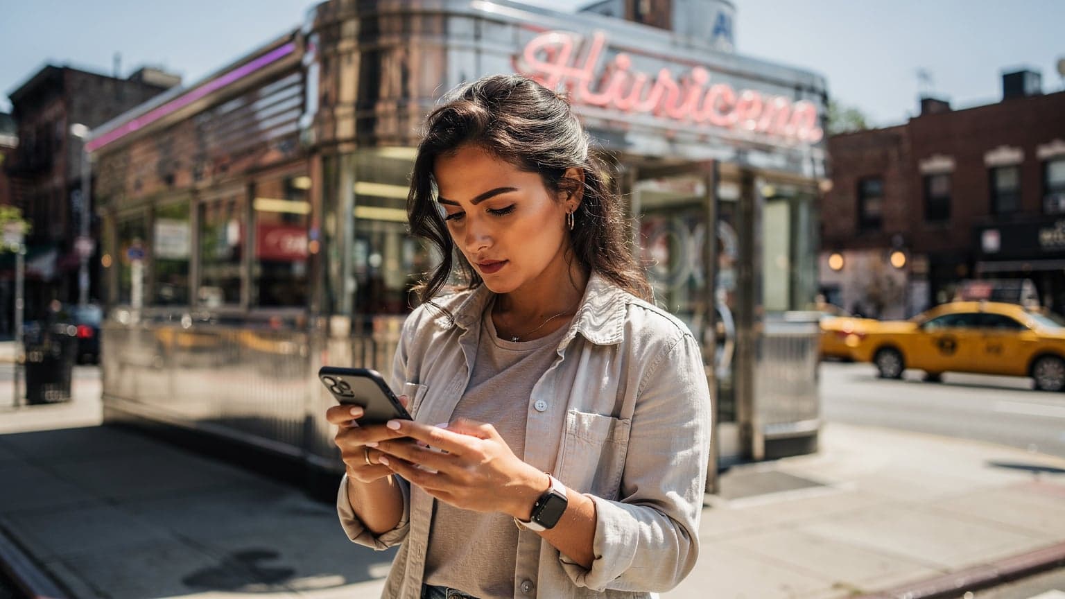 Una viaggiatrice italiana consulta il suo smartphone con un'espressione decisa mentre si trova all'esterno di un tipico diner americano a Brooklyn, New York, in una giornata soleggiata.