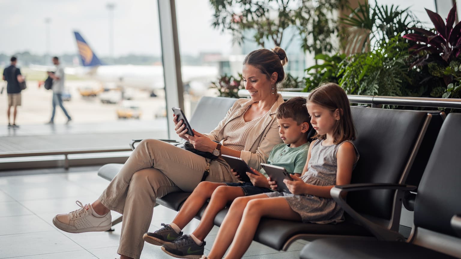 Seorang ibu tersenyum lega sambil memegang smartphone-nya di tengah keramaian bandara Changi Singapura, dengan anak-anaknya yang fokus bermain tablet di sampingnya.