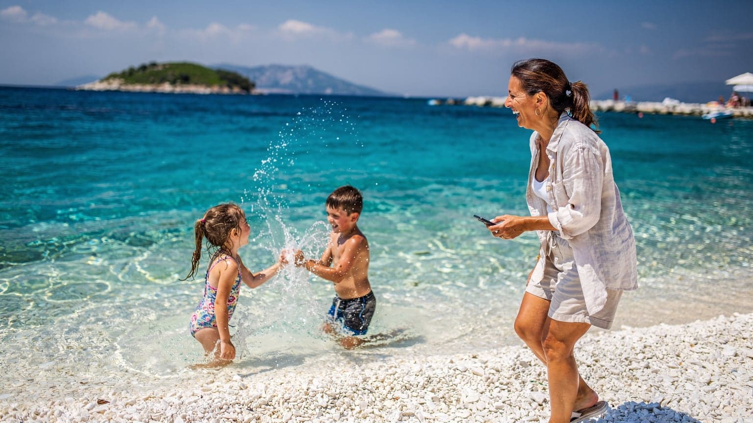 Μια μητέρα με τα δύο παιδιά της, περίπου 8 και 10 ετών, χαμογελούν στην παραλία των Αγίων Σαράντα στην Αλβανία, με τα κρυστάλλινα γαλάζια νερά και τον καταγάλανο ουρανό στο παρασκήνιο. Η μητέρα κρατάει ένα smartphone με μια θήκη σε παστέλ χρώμα.