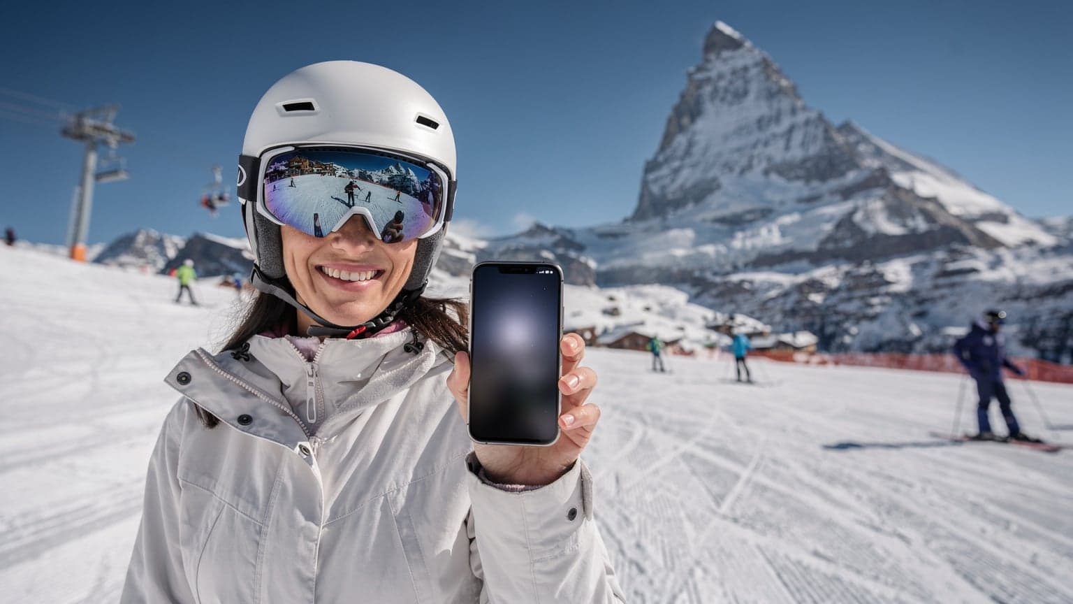 Eine Frau in Skibekleidung lächelt und hält ihr Smartphone hoch, während sie auf einer sonnigen Skipiste in Zermatt, Schweiz, steht, mit dem Matterhorn im Hintergrund.