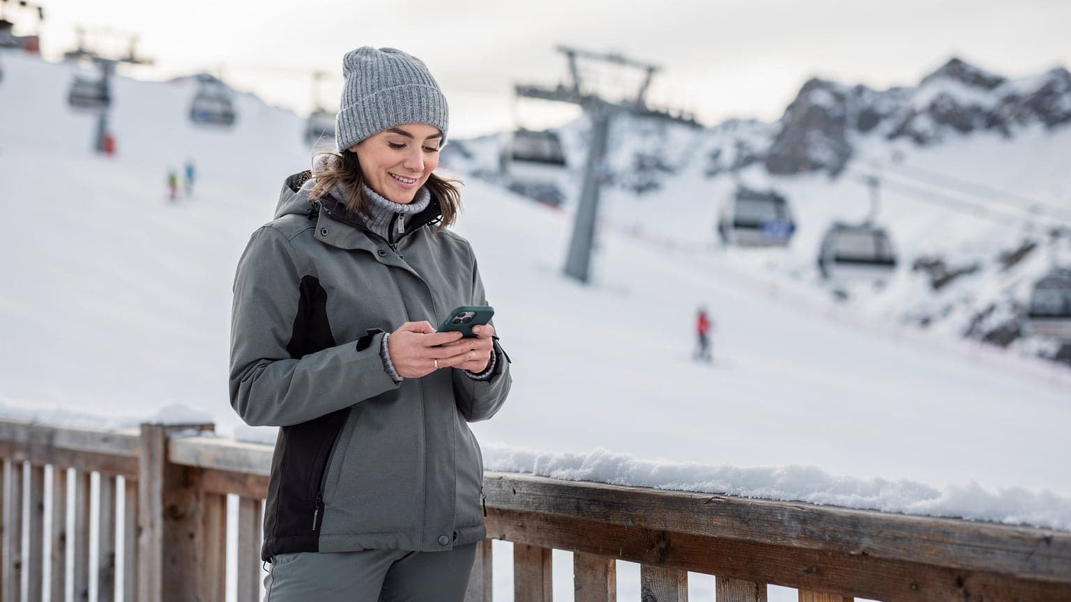 Una mujer con gorro de lana y chaqueta de esquí, sonríe mientras revisa su teléfono en una estación de esquí en Andorra, con montañas nevadas de fondo y un telecabina pasando por encima.