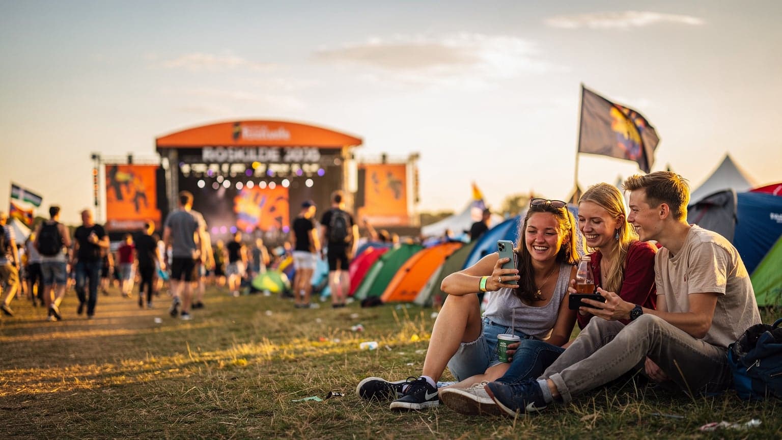 En ung kvinde navigerer sin smartphone gennem et tætpakket festivalområde i skumringen, med scener og telte i baggrunden, tydeligt optaget af at finde vej eller dele et øjeblik.
