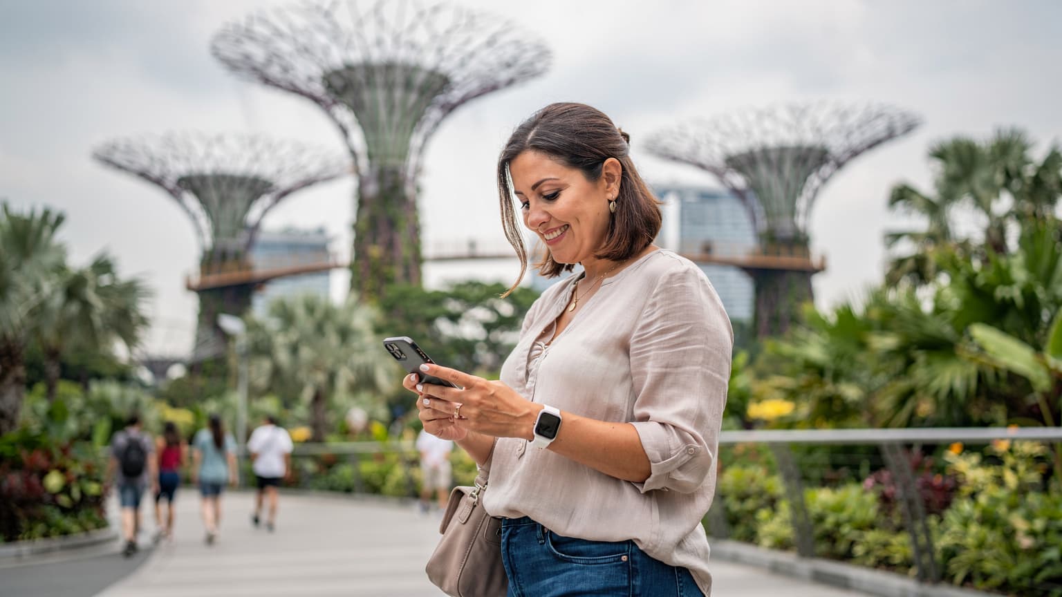 Seorang ibu tersenyum sambil memegang ponselnya di Gardens by the Bay, Singapura, dengan latar belakang Supertrees yang ikonik.