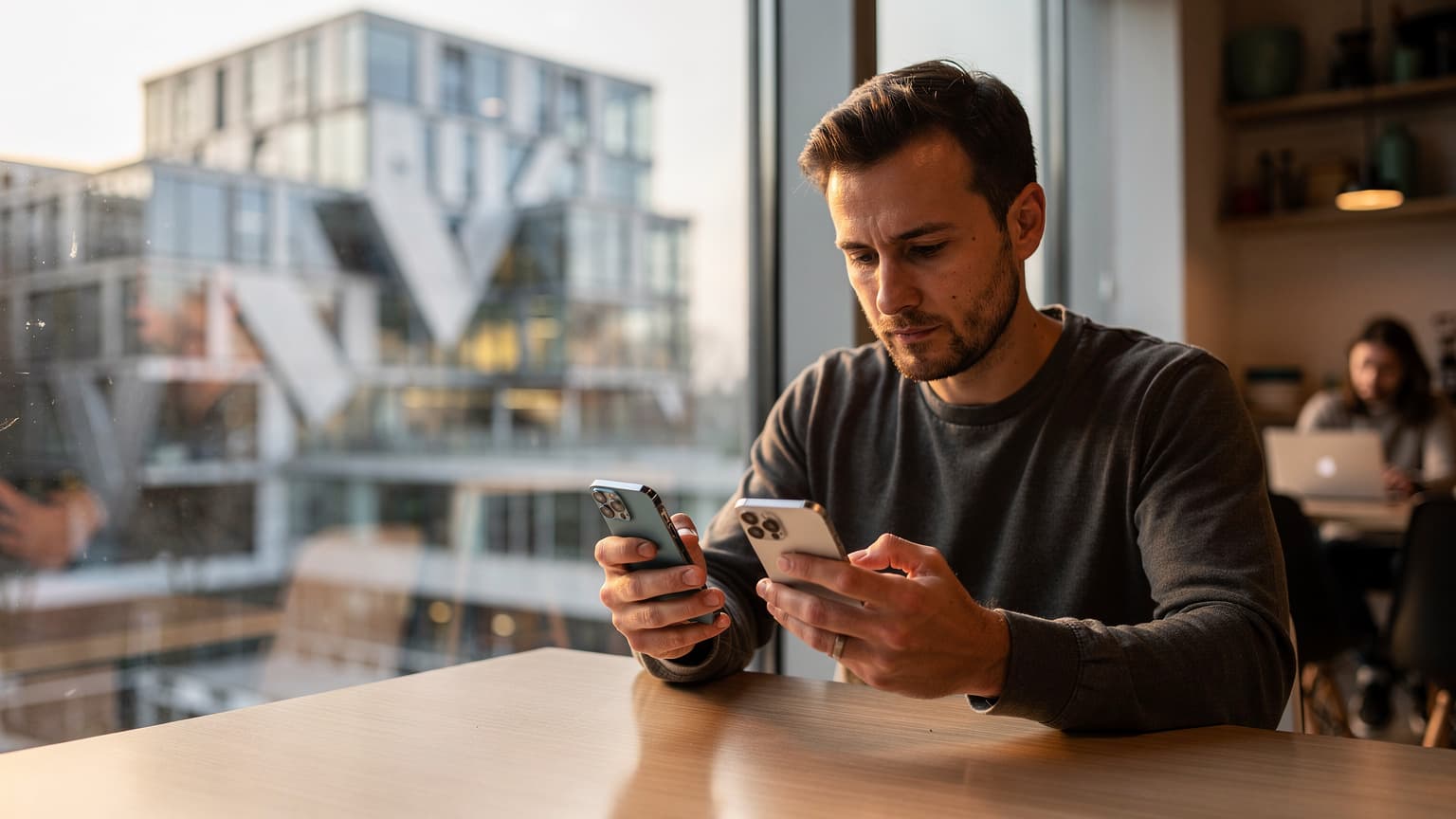 Un homme d'une trentaine d'années, l'air concentré, transférant son eSIM d'un iPhone à un autre, assis à une table dans un café moderne et lumineux du quartier de La Confluence à Lyon, la lumière du soleil filtrant à travers une grande baie vitrée.