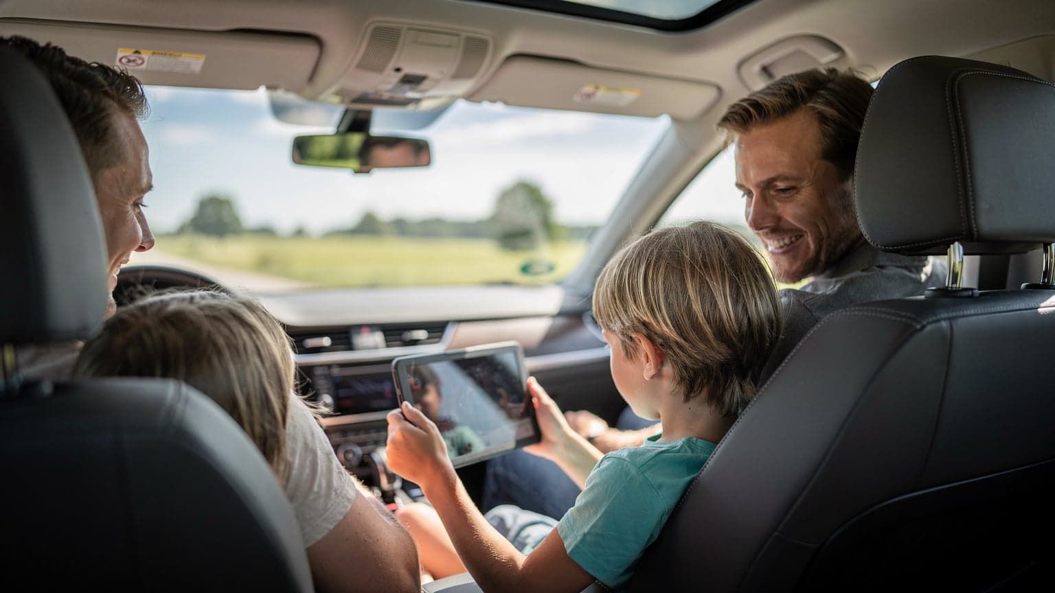En familie på fire, mor, far og to børn, sidder i en bil på vej mod en dansk feriedestination, med udsigt til grønne marker og en blå sommerhimmel, mens et barn holder en tablet og ser en film.