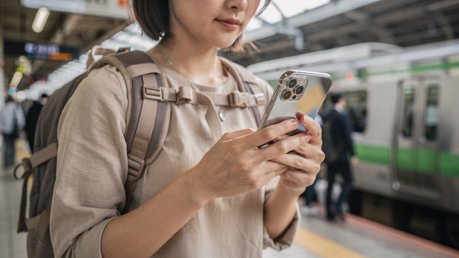 東京の新宿駅周辺で、iPhoneを使ってeSIM設定を確認している旅行者の手元が写っている。背景には、電車が発着するプラットフォームと、高層ビル群がぼんやりと見える。