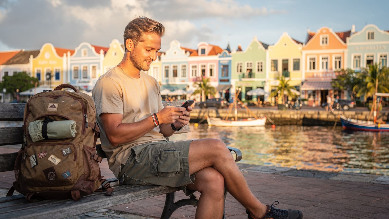 Een jonge backpacker zit ontspannen op een bankje in de haven van Willemstad, Curaçao, met een telefoon in de hand en de iconische pastelkleurige gebouwen van de Handelskade op de achtergrond.