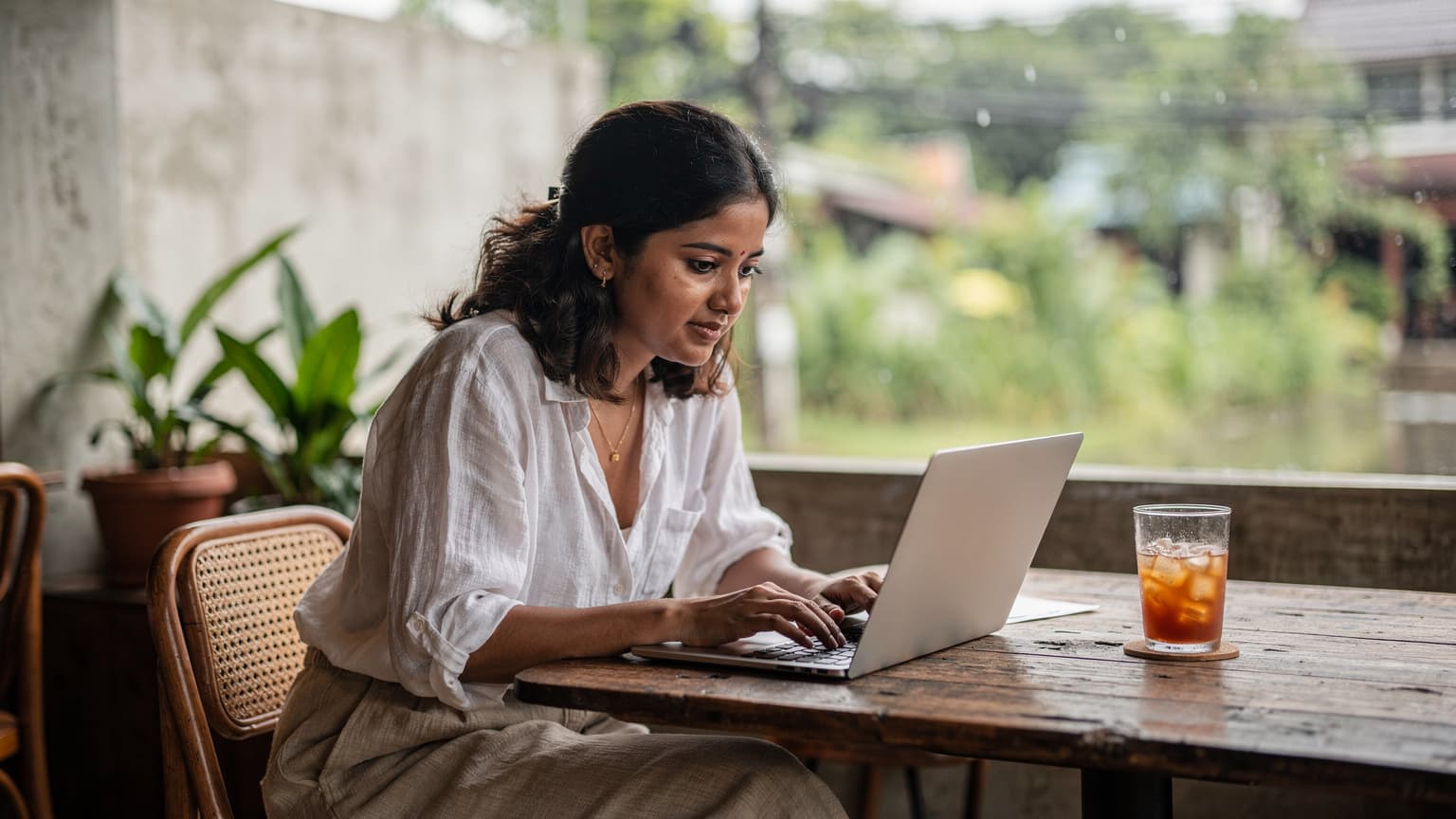 थाईलैंड के चियांग माई में एक कैफे में बैठी एक युवा भारतीय महिला लैपटॉप पर काम कर रही है, उसके पास एक ग्लास आइस्ड थाई टी है और वह बारिश से भरे हरे-भरे परिदृश्य को देख रही है.
