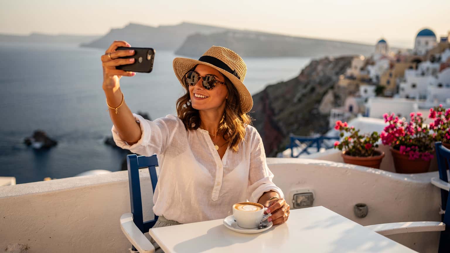 Una donna sorridente, con occhiali da sole e un cappello di paglia, tiene in mano il suo smartphone mentre si gode un caffè su una terrazza panoramica a Santorini, Grecia, con il Mar Egeo e le iconiche case bianche sullo sfondo.