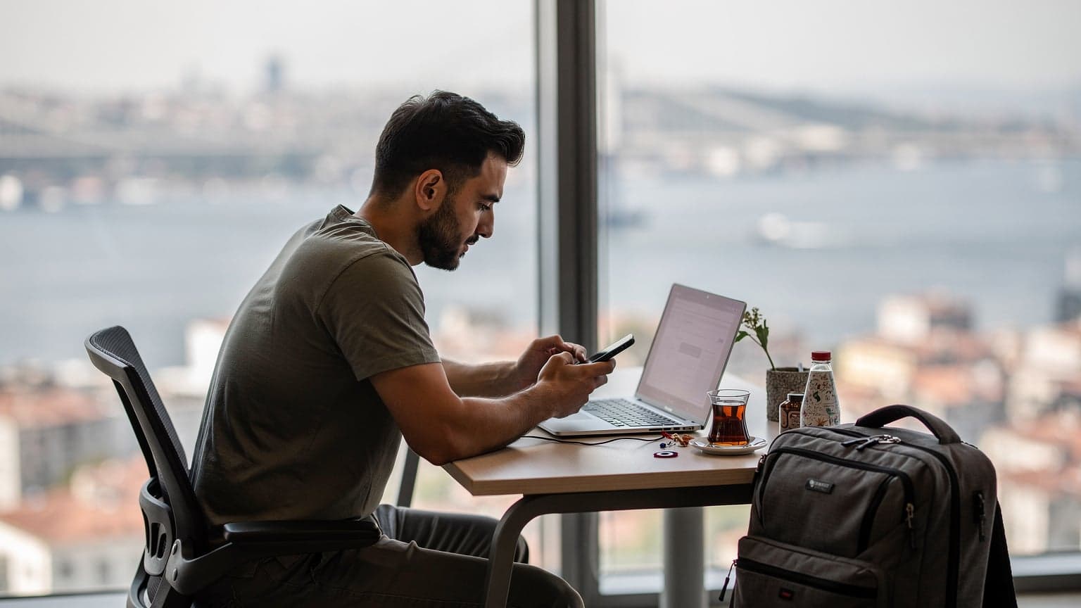 İstanbul'daki bir co-working space'te laptopuyla çalışan bir dijital göçebe, elinde akıllı telefon ve bir kahve bardağı tutuyor, arka planda Boğaz Köprüsü'nün silüeti hafifçe görünüyor.