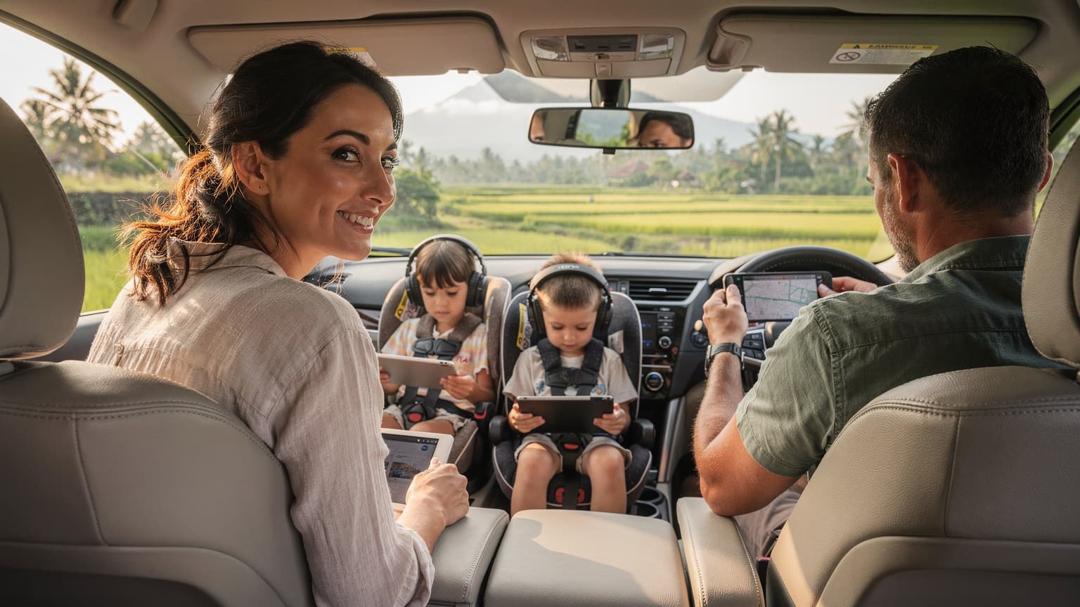 Seorang ibu tersenyum lega di samping suaminya yang sedang membantu anak-anak mereka memasang headphone di kursi belakang mobil, dengan pemandangan pegunungan Bali yang hijau di luar jendela mobil, menunjukkan momen perjalanan keluarga yang tenang dan terhubung.