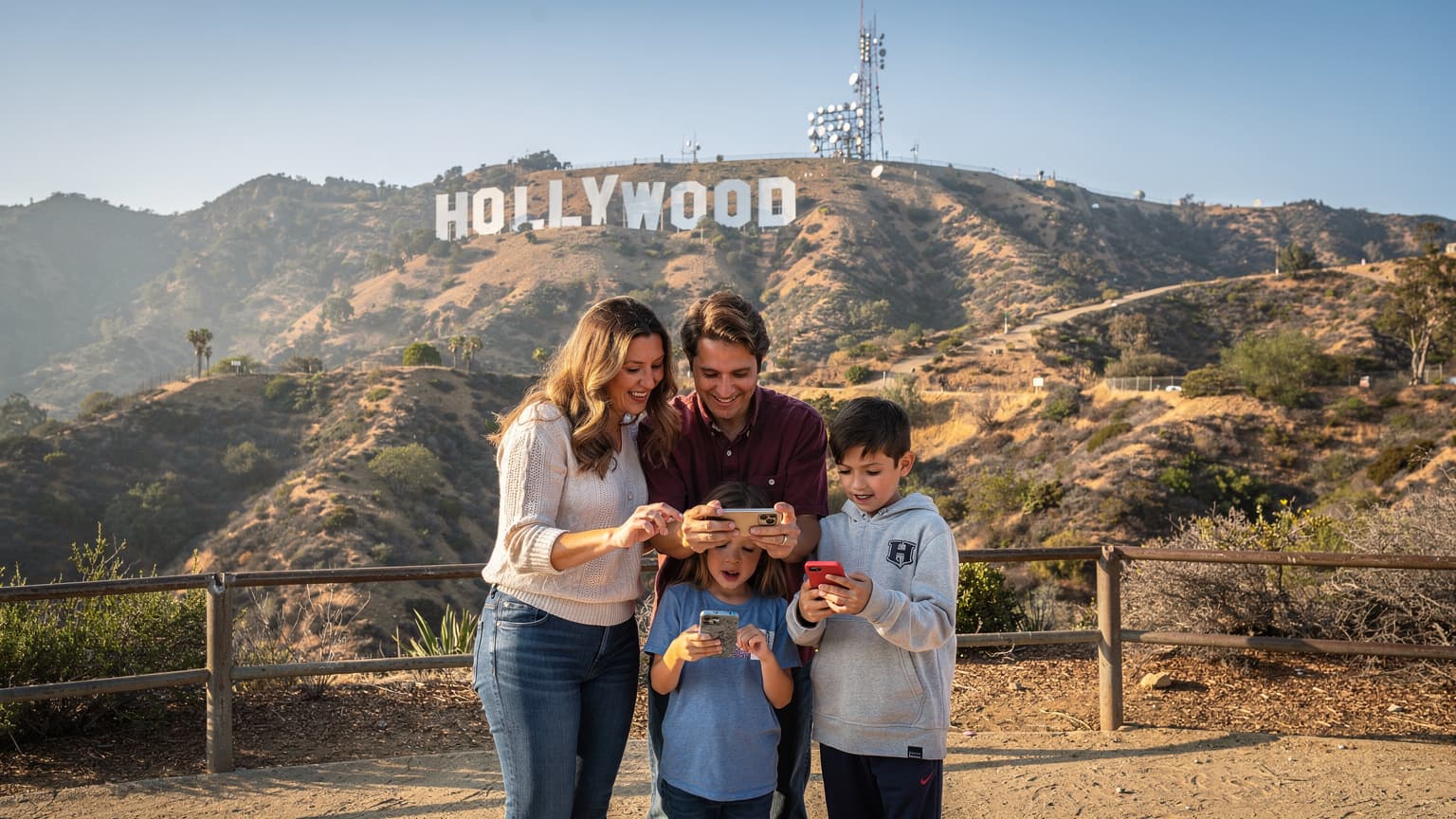 Una familia, dos adultos y dos niños, emocionados usando sus teléfonos para navegar mientras posan frente al icónico letrero de Hollywood en Los Ángeles bajo un cielo azul brillante.