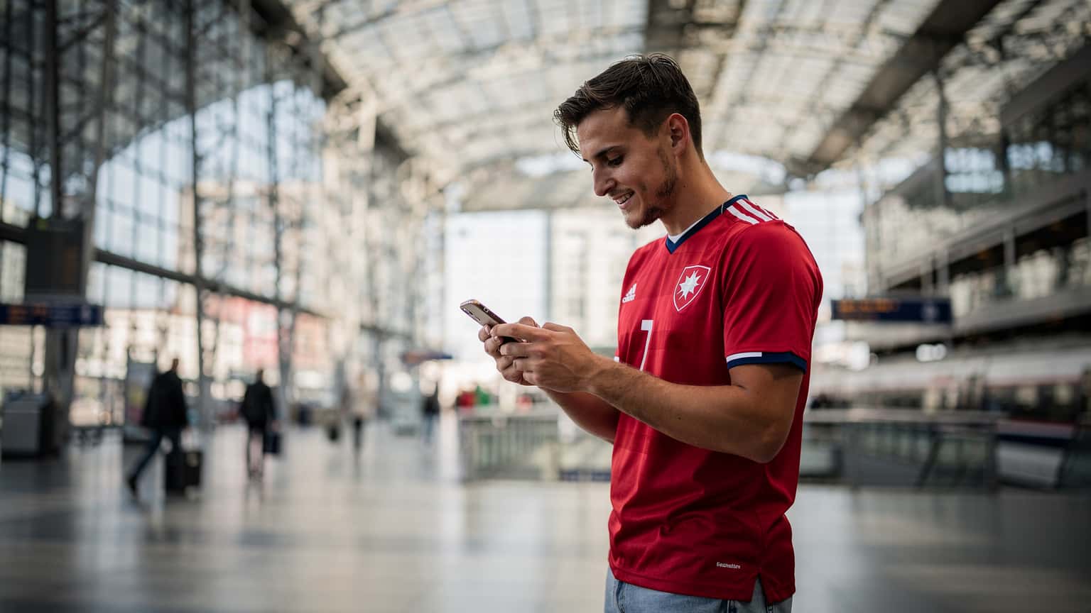 Mladý muž v dresu české fotbalové reprezentace s chytrým telefonem v ruce kontroluje výsledky zápasů Euro 2026 na hlavním nádraží v Berlíně, s moderní architekturou v pozadí.