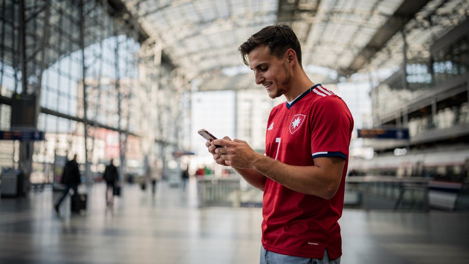 Mladý muž v dresu české fotbalové reprezentace s chytrým telefonem v ruce kontroluje výsledky zápasů Euro 2026 na hlavním nádraží v Berlíně, s moderní architekturou v pozadí.