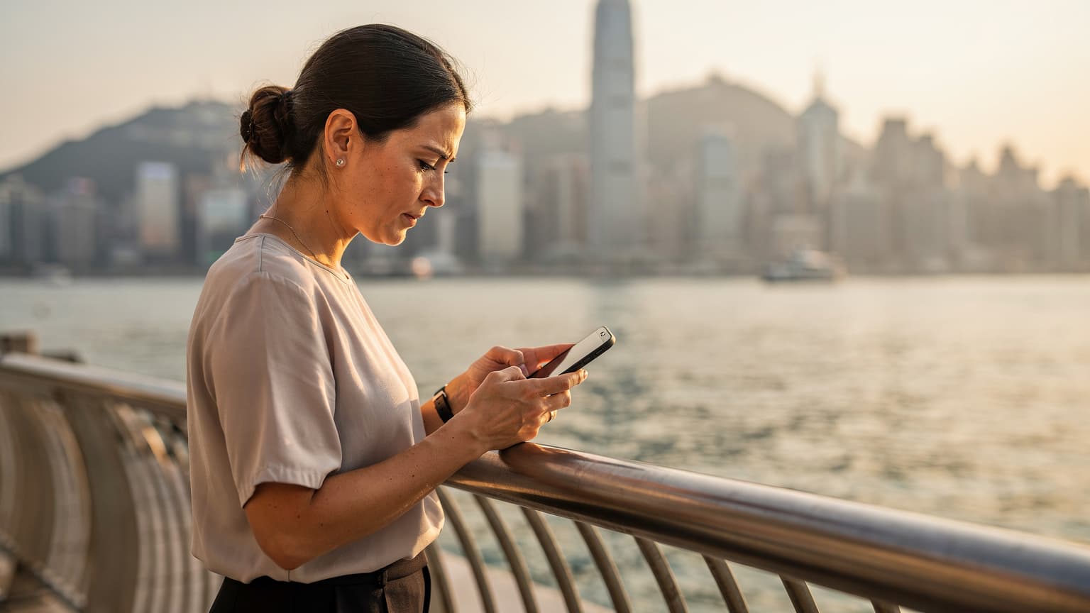 一位專業的電信工程師在香港維多利亞港邊,手持智慧手機,仔細查看eSIM網路連接狀況的畫面。