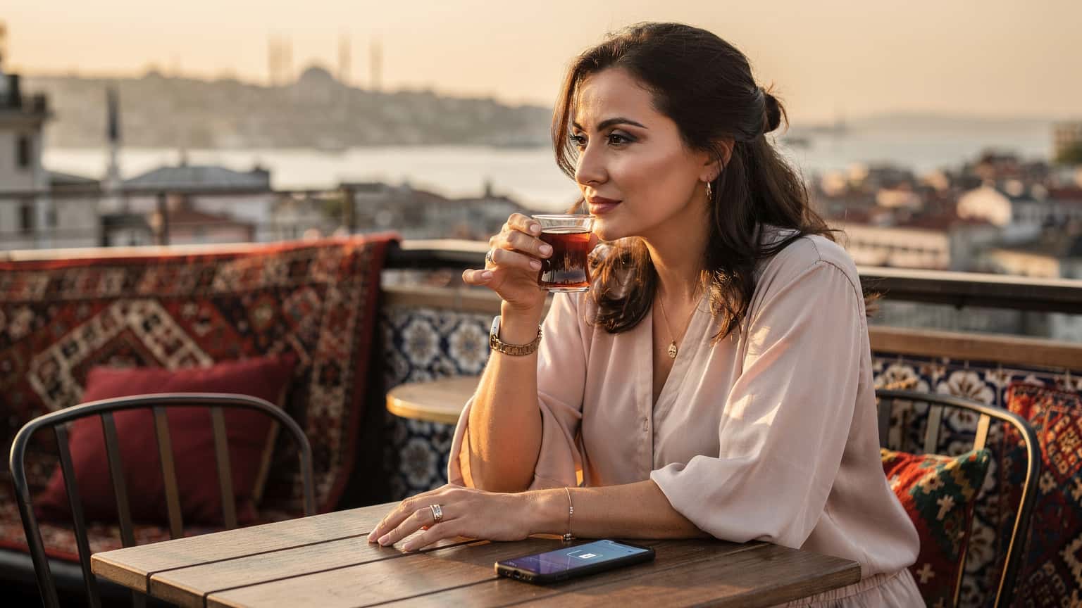 Eine Frau trinkt türkischen Tee auf einer Dachterrasse in Beyoğlu, Istanbul, mit Blick auf den Bosporus bei Sonnenuntergang und hält diskret ein Smartphone in der Hand, das eine Cellesim eSIM anzeigt.