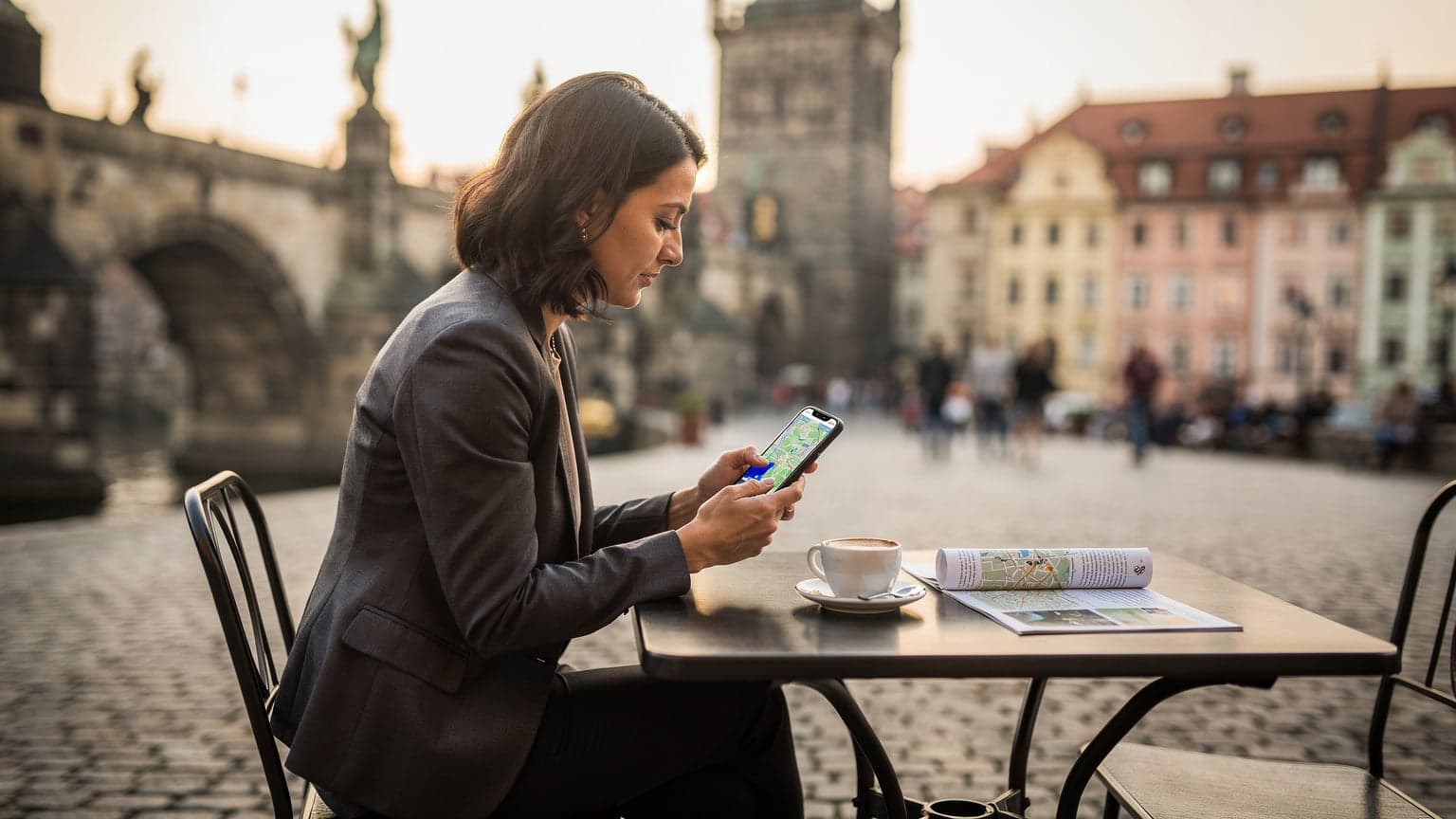 Žena sedící v kavárně v Malé Straně v Praze, dívající se na svůj telefon s mapou, s Karlovým mostem v pozadí.