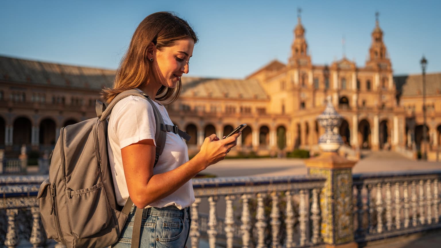 Una viajera, con una mochila ligera y un sombrero de ala ancha, revisa su teléfono en la Plaza de España de Sevilla, con la arquitectura ornamentada de fondo, buscando indicaciones para su próximo destino.