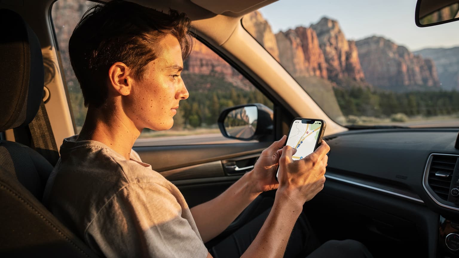 A person in a passenger seat of a car, holding a smartphone with a map application open, looking at the road ahead during a sunset drive through a scenic American landscape.