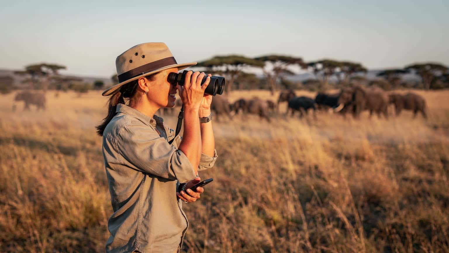 Een vrouw met een zonnehoed kijkt door een verrekijker naar een kudde olifanten in de verte, terwijl ze een smartphone met een eSIM in haar hand houdt, met de uitgestrekte savanne van het Krugerpark op de achtergrond tijdens zonsondergang.