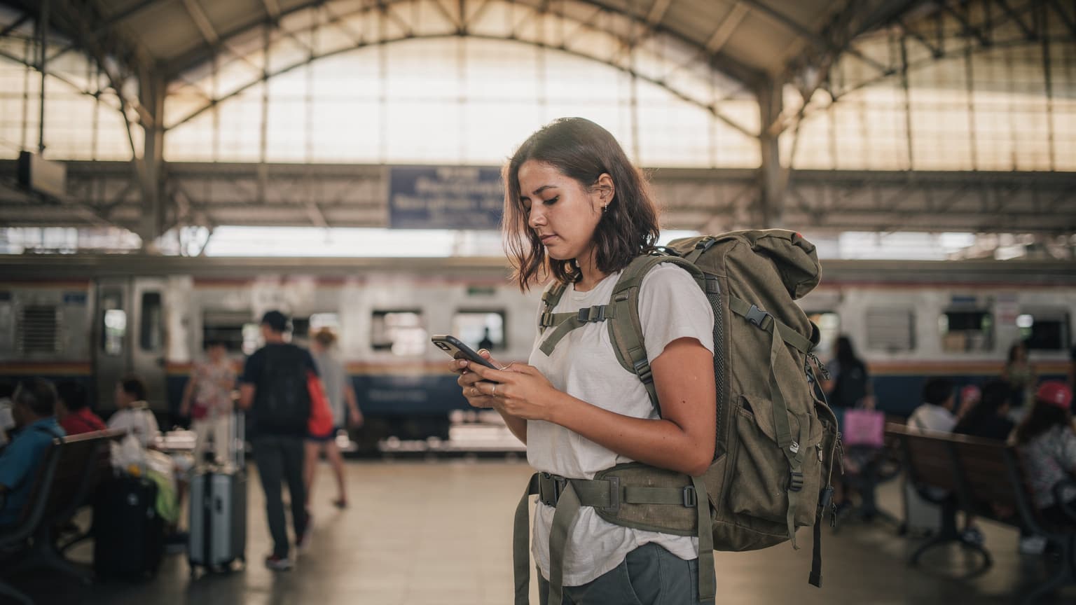 A budget backpacker in a busy train station in Bangkok, Thailand, looking at her iPhone screen with a focused expression, checking her eSIM details before boarding a train.