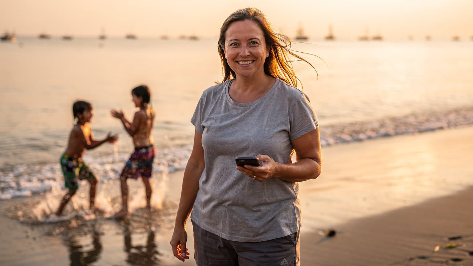 Seorang ibu tersenyum sambil memegang ponselnya di pinggir pantai Sanur, Bali, dengan anak-anak bermain di latar belakang saat matahari terbit.