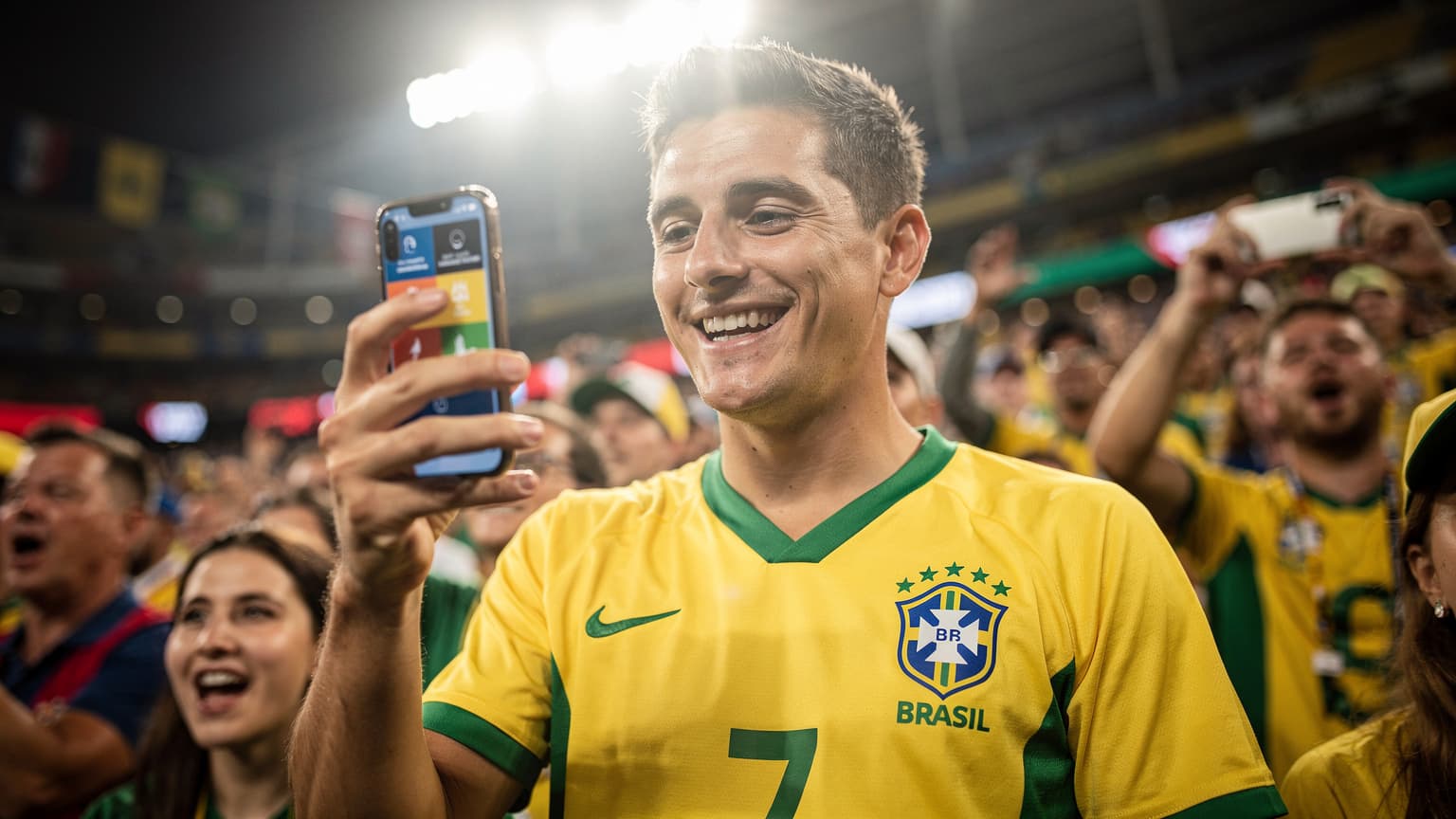 Uma pessoa animada em um estádio lotado nos EUA, com a camisa da seleção brasileira, segurando um smartphone com um sorriso enquanto verifica a conexão, com a tela mostrando um aplicativo de placares de futebol.