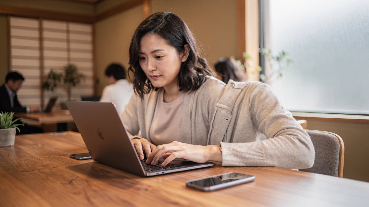 A digital nomad sits at a wooden table in a busy Tokyo co-working space, focused on her laptop with a smartphone next to it, bathed in warm morning light streaming through a large window.