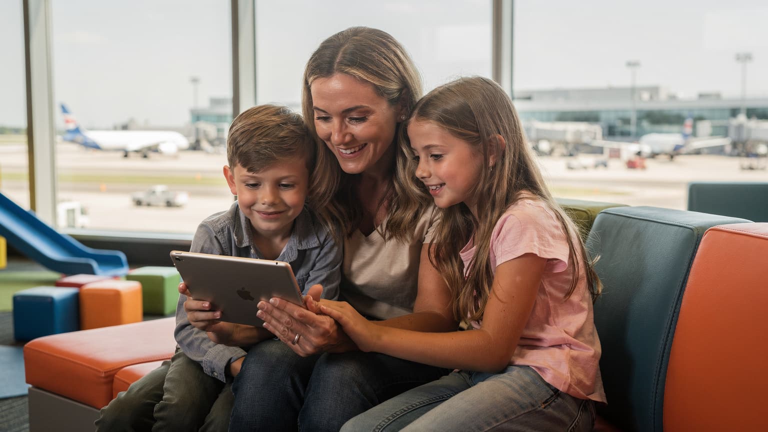 Una madre e due bambini, circa di 8 e 10 anni, sorridono mentre guardano un tablet in un'area giochi di un aeroporto affollato negli Stati Uniti, con finestre che mostrano aerei in lontananza.
