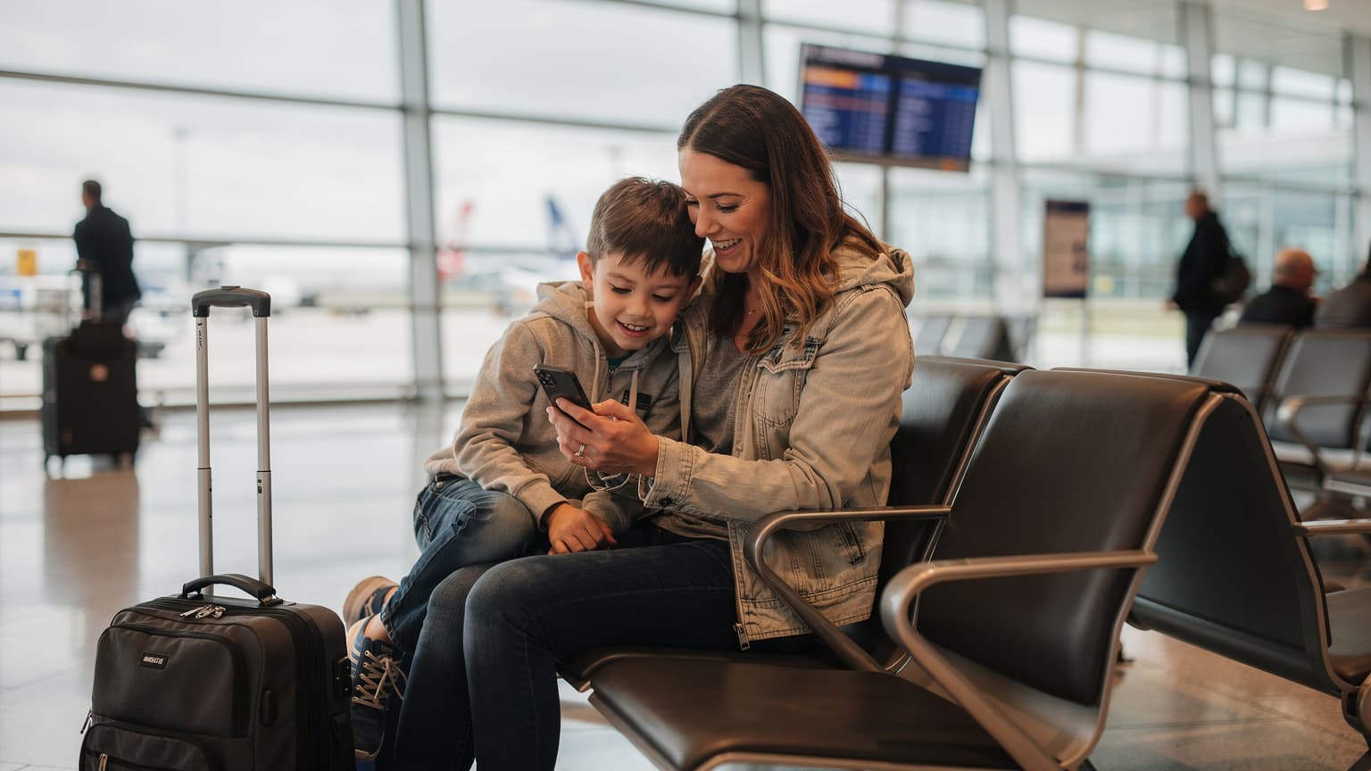 Une mère souriante et son fils regardent attentivement un iPhone, assis confortablement dans la salle d'attente animée de l'aéroport Roissy-Charles de Gaulle, avec des valises à roulettes à leurs côtés.