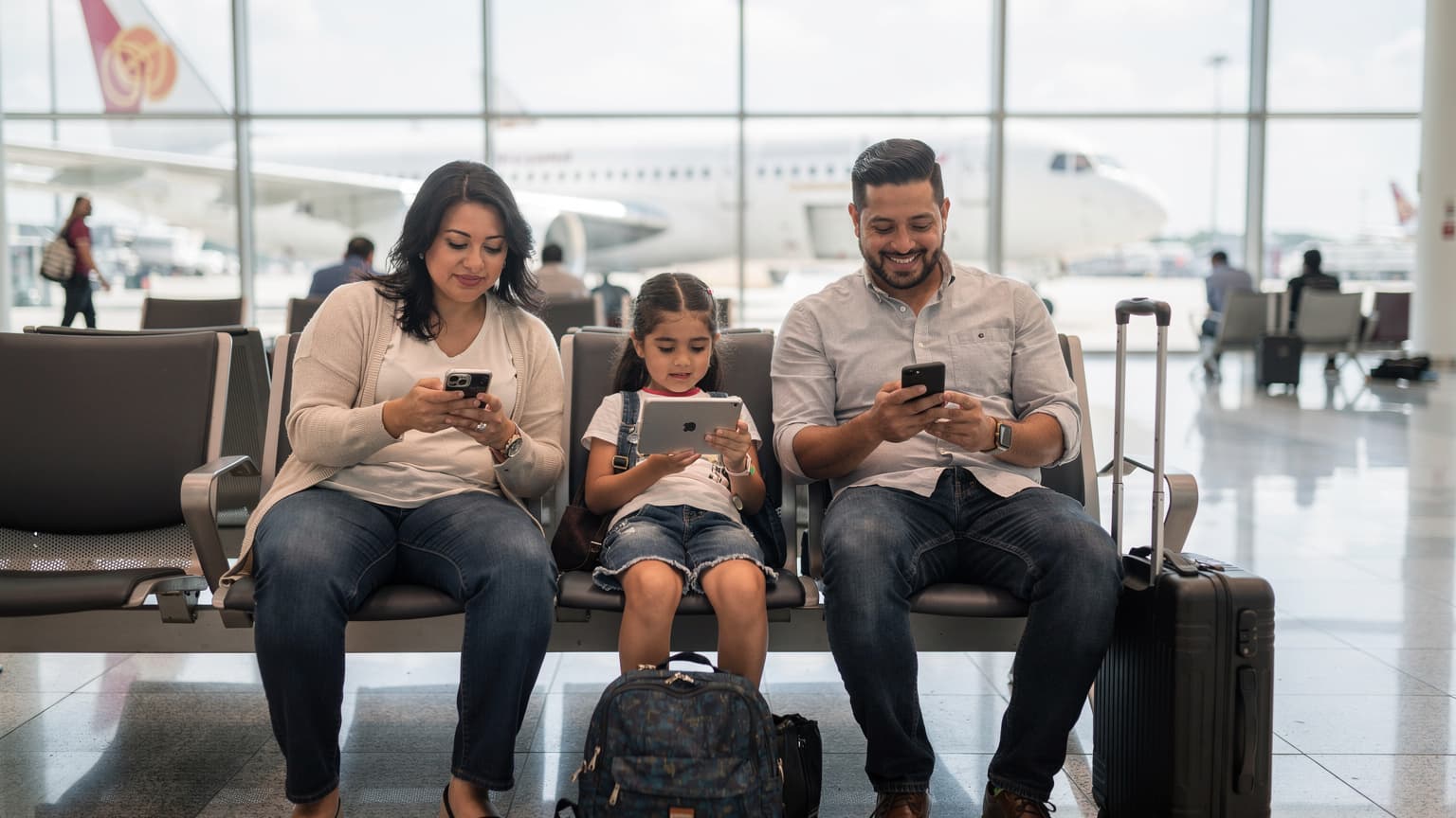 Una familia hispana, padres y dos niños de 8 y 10 años, en el aeropuerto Changi de Singapur, con los niños entretenidos en sus iPads y los padres sonriendo mientras revisan sus teléfonos.