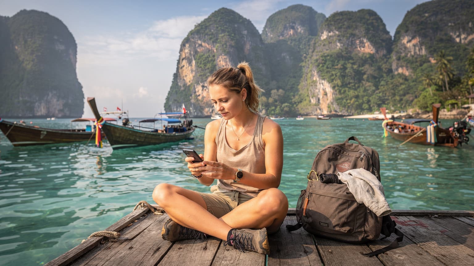 Młoda podróżniczka z plecakiem sprawdza telefon z eSIM, siedząc na drewnianym pomoście na tle turkusowej wody i łodzi longtail na Koh Phi Phi w Tajlandii.