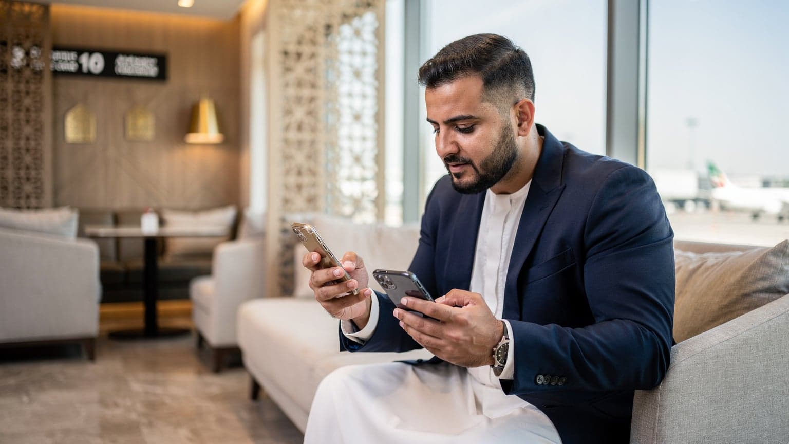 شخص يحمل هاتفين ذكيين, أحدهما جديد والآخر قديم, في صالة انتظار حديثة ومشرقة بمطار دبي الدولي, مع حقيبة سفر أنيقة بجانبه.
