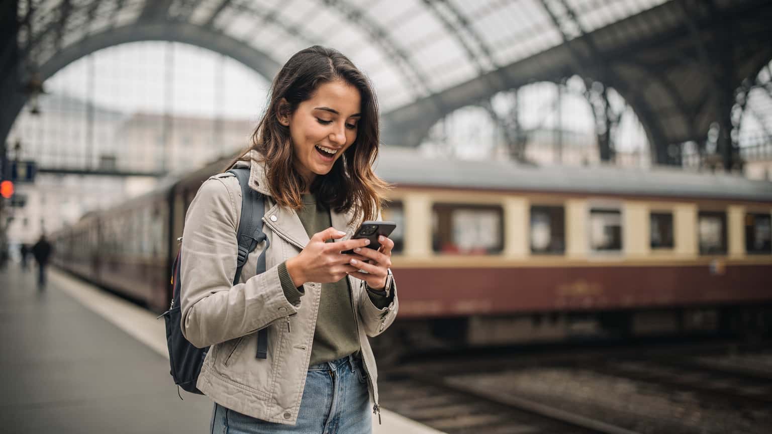 A woman excitedly checking her phone at a bustling European train station, with a vintage train visible in the background, showing immediate connectivity.