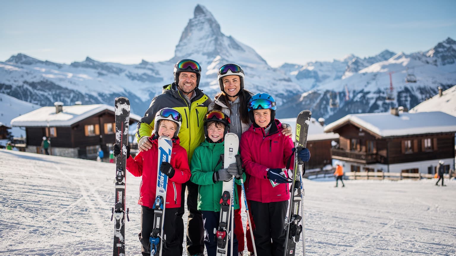 Uma família brasileira, pais e duas crianças de 8 e 10 anos, sorriem enquanto posam para uma foto com esquis e bastões em uma pista de esqui nevada nos Alpes suíços, com chalés de madeira e picos cobertos de neve ao fundo.