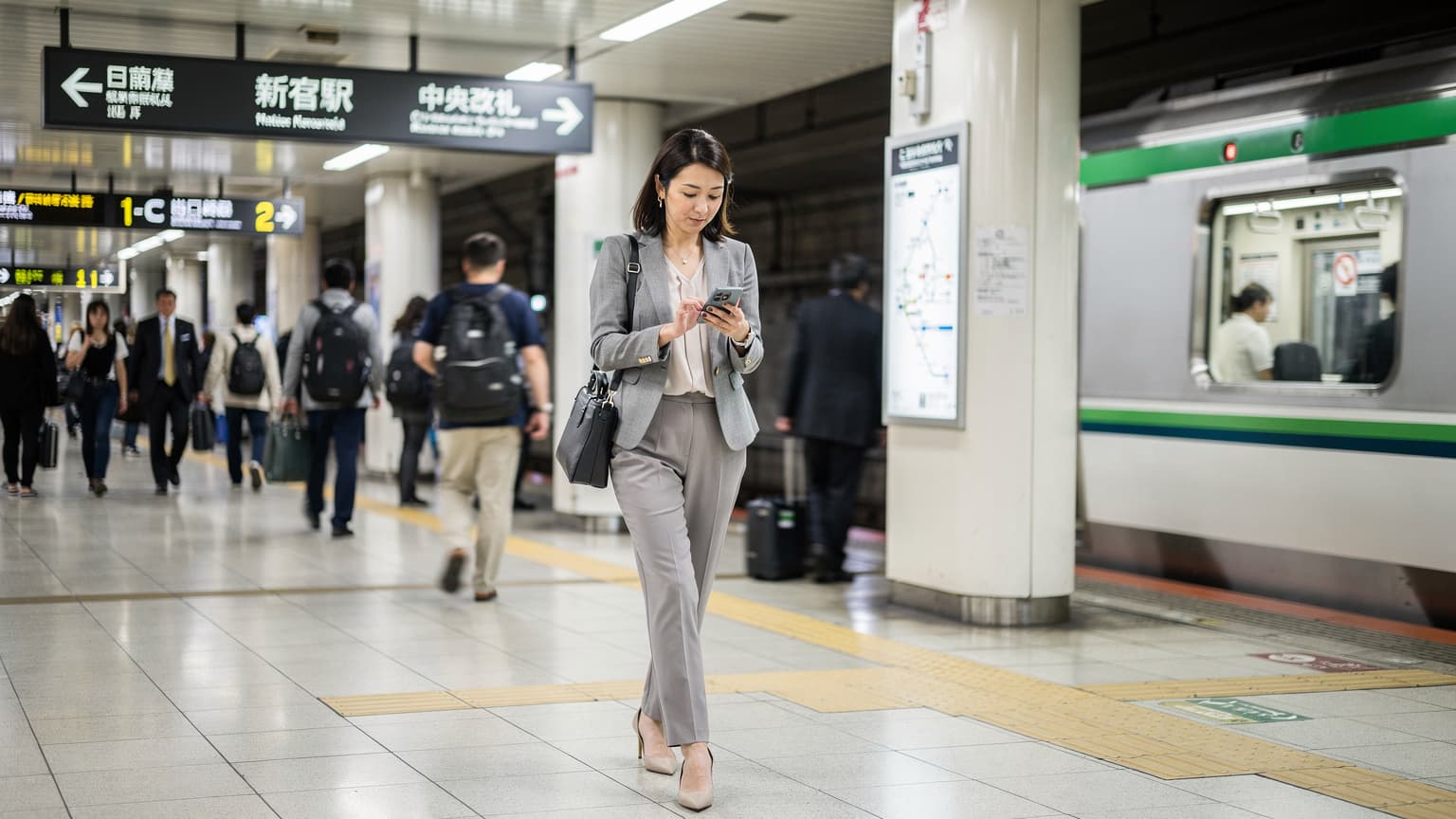A woman confidently navigating a busy train station in Tokyo, holding her smartphone, with a JR East ticket gate in the background, suggesting seamless connectivity.