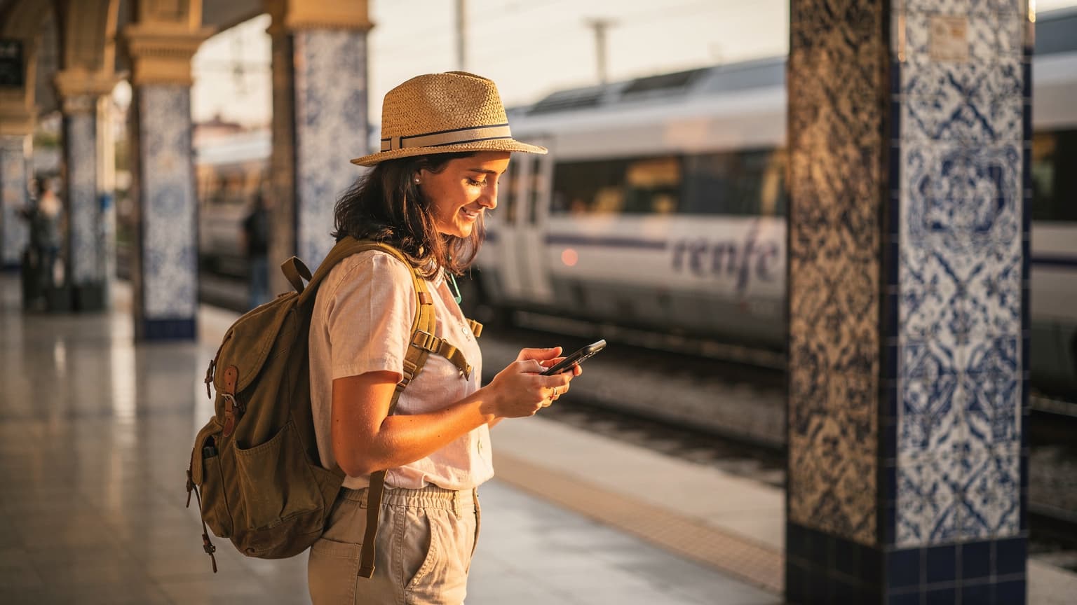 Una viajera joven y sonriente, con mochila y sombrero de paja, consulta su teléfono móvil mientras espera un tren en la estación de Santa Justa en Sevilla, España, con los azulejos andaluces de fondo.