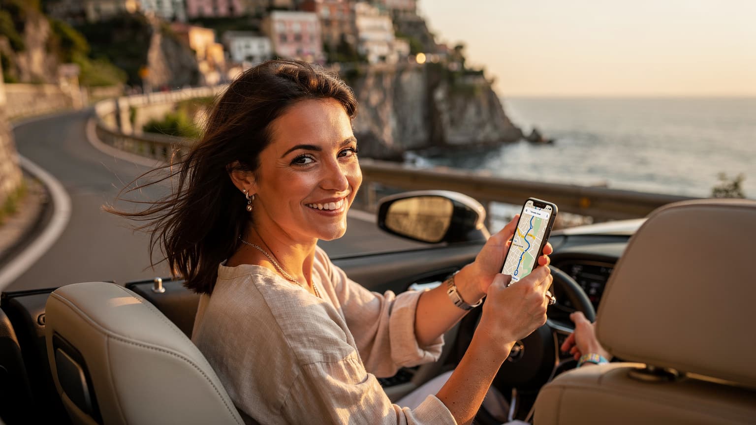 Une femme souriante, les cheveux au vent, tient son smartphone avec une carte de navigation affichée, assise sur le siège passager d'une voiture décapotable, traversant une route côtière en Italie au coucher du soleil.