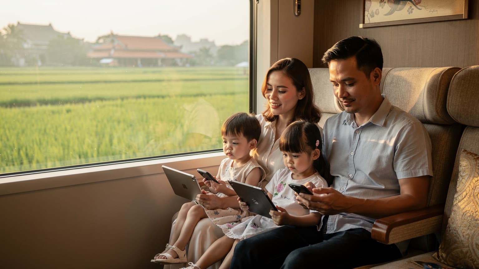 Một gia đình Việt Nam với bố, mẹ và hai con nhỏ đang ngồi thoải mái trên tàu hỏa cao cấp nhìn ngắm khung cảnh đồng lúa xanh mướt chạy qua cửa sổ, với iPad và điện thoại trên tay bọn trẻ, chụp theo phong cách ảnh bìa tạp chí du lịch.