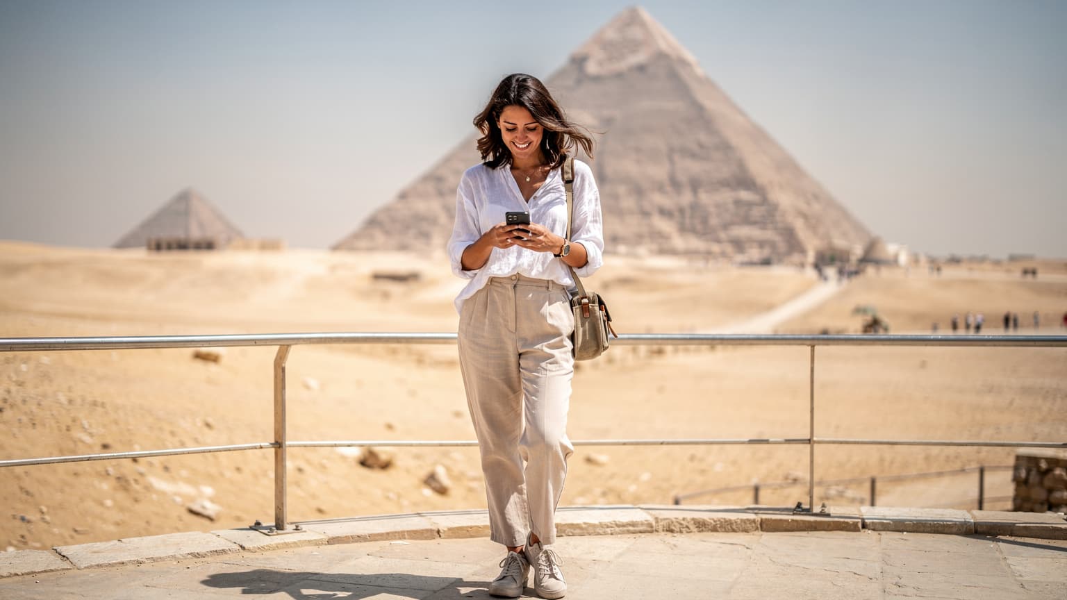 Una viaggiatrice sorride mentre naviga sul suo smartphone, con lo sfondo sfocato delle Piramidi di Giza sotto il cielo blu intenso.