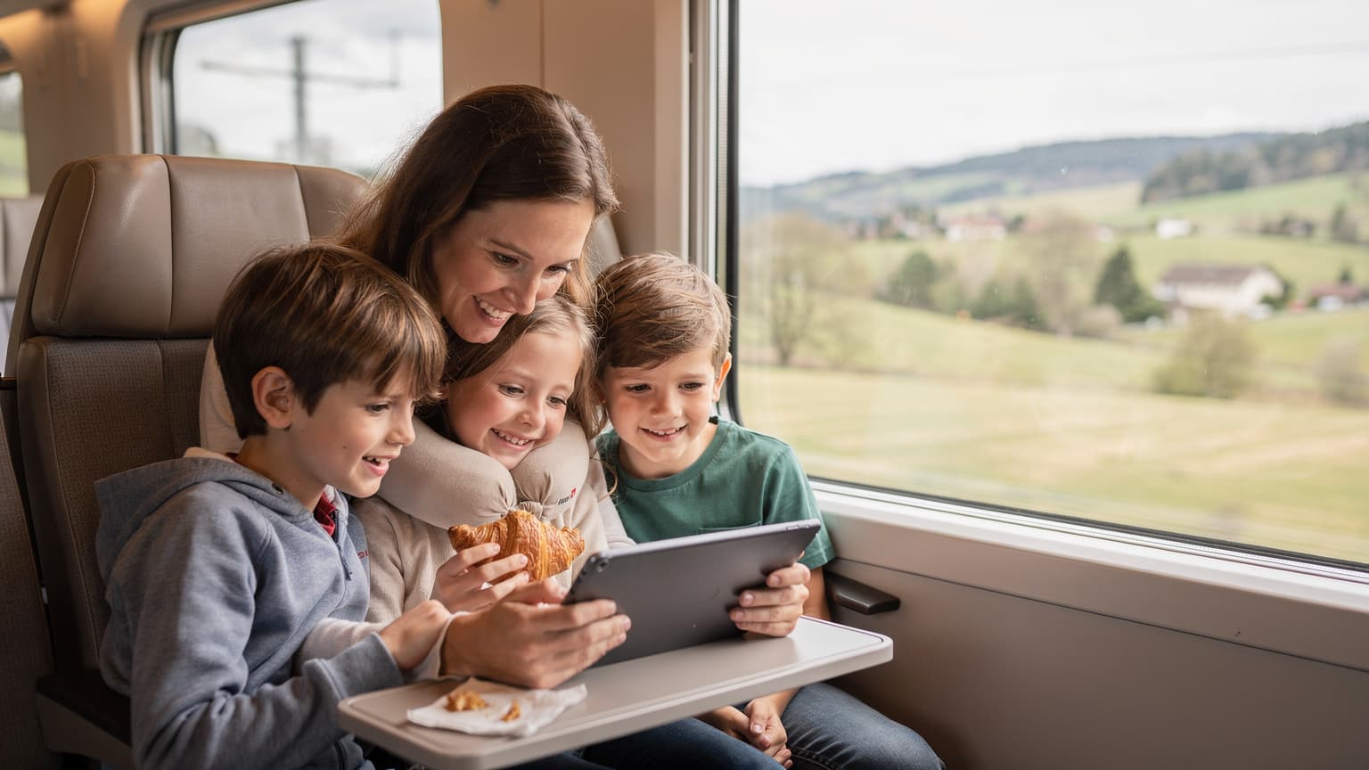 A smiling mother and her two children, aged around 8 and 10, are looking at a tablet together on a comfortable train seat, with a scenic European landscape visible through the window.