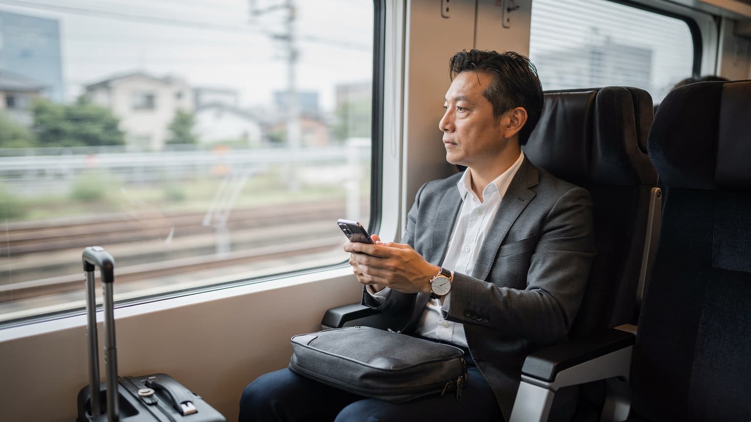 東京駅から成田空港へ向かう電車内で、窓の外を眺めながらスマートフォンでeSIM設定を最終確認するビジネスパーソン。