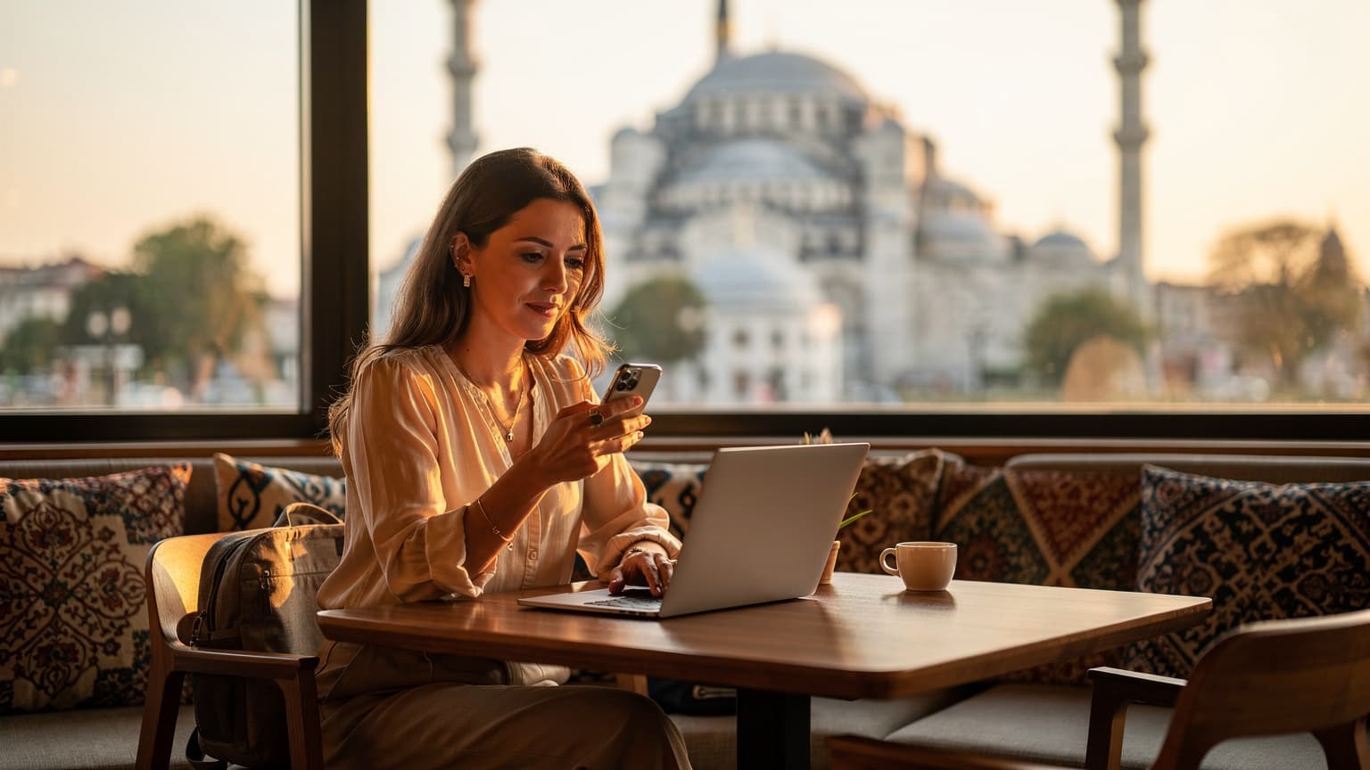İstanbul'da, Sultanahmet Camii manzaralı bir kafede laptopunda çalışan bir dijital göçebe kadın, elinde akıllı telefonuyla eSIM kurulumu yaparken gülümsüyor.