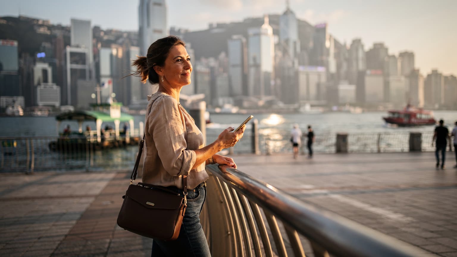 一名旅客在香港繁忙的尖沙咀海濱,手持智能手機,背景是維多利亞港和香港島的摩天大樓,夕陽餘暉灑落。