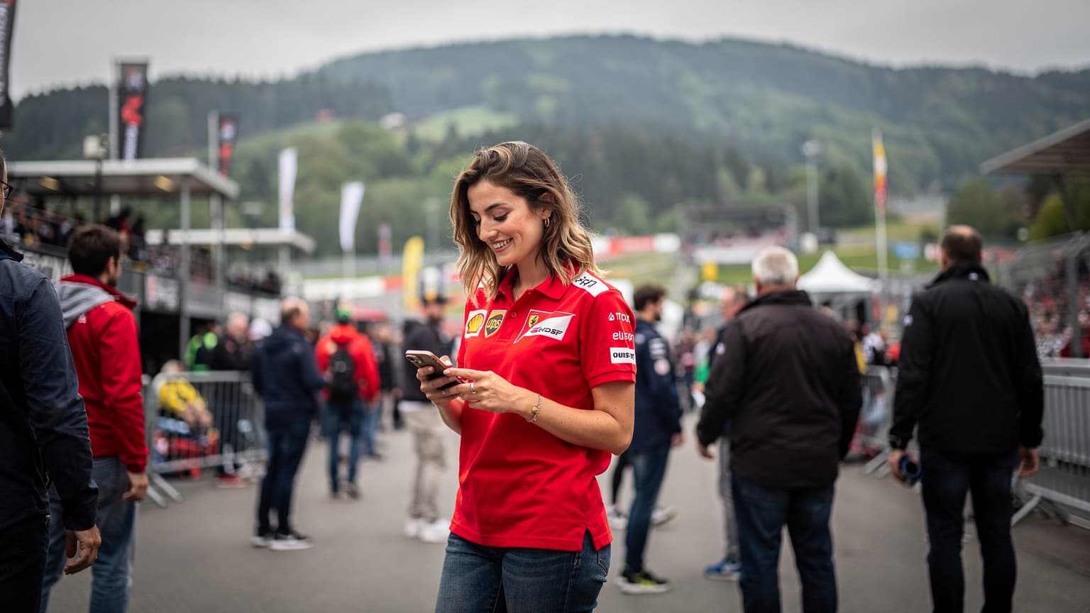 Een jonge vrouw in een rode race-shirt met een smartphone in haar hand, lachend terwijl ze door de drukte van Spa-Francorchamps navigeert, met de iconische Ardennenheuvels op de achtergrond.
