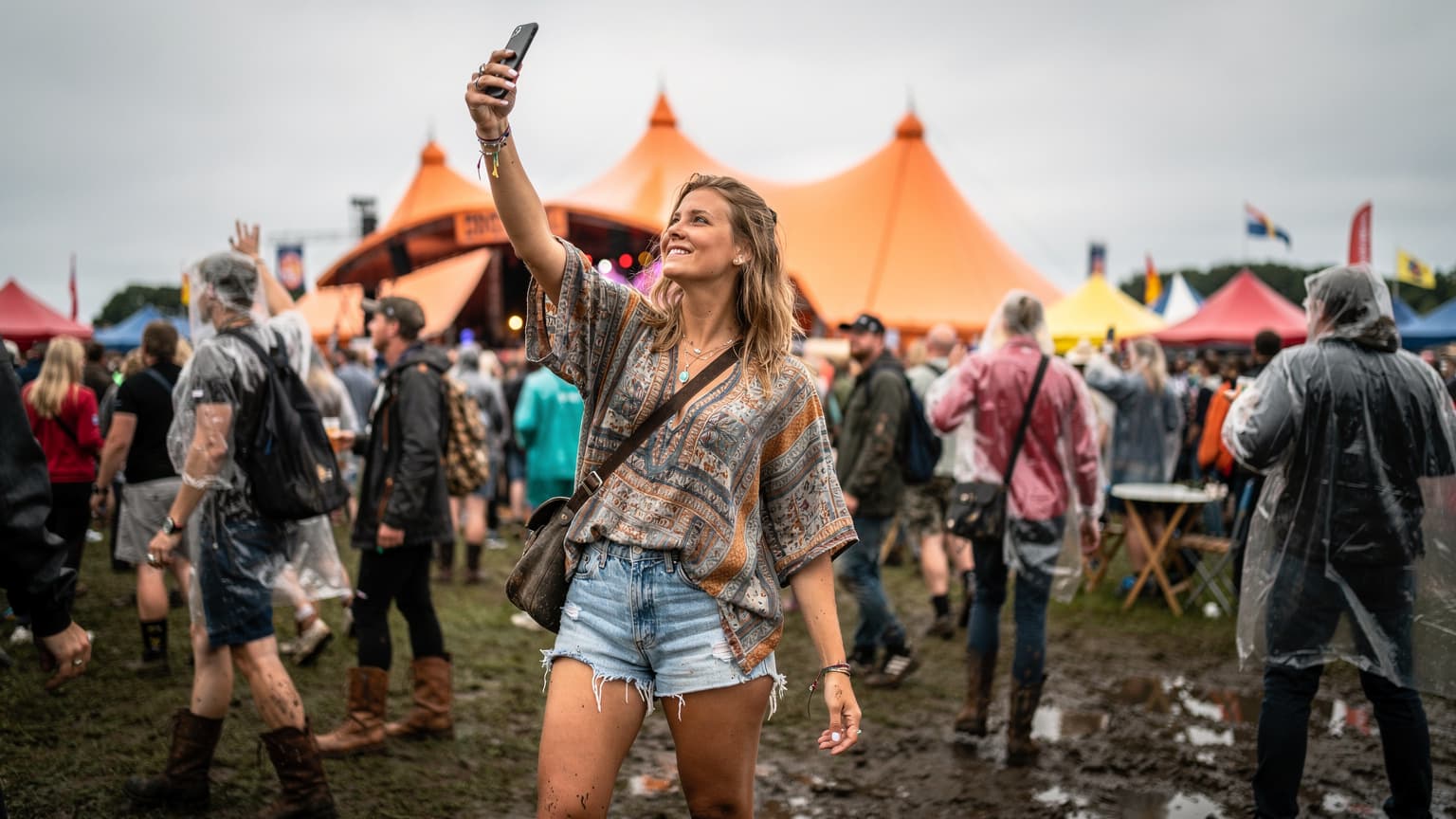 En ung kvinde i festivalklæder smiler og holder sin smartphone op i luften for at fange et signal midt i en stor folkemængde på Roskilde Festivalens orange scene, med mudder på hendes støvler og farverige telte i baggrunden under en overskyet sommerhimmel.